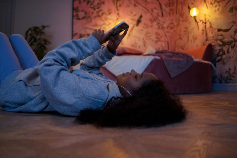 A young woman relaxes while browsing her smartphone in a cozy, softly lit bedroom at night.